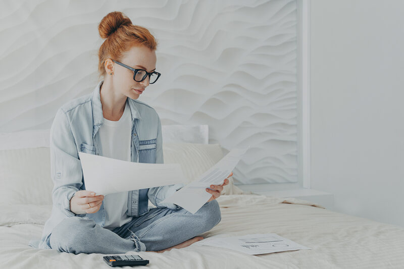 Young concentrated woman housewife in spectacles managing family budget while sitting on bed at home, redhead female calculating bills household expenses, holding papers with charts and graphics